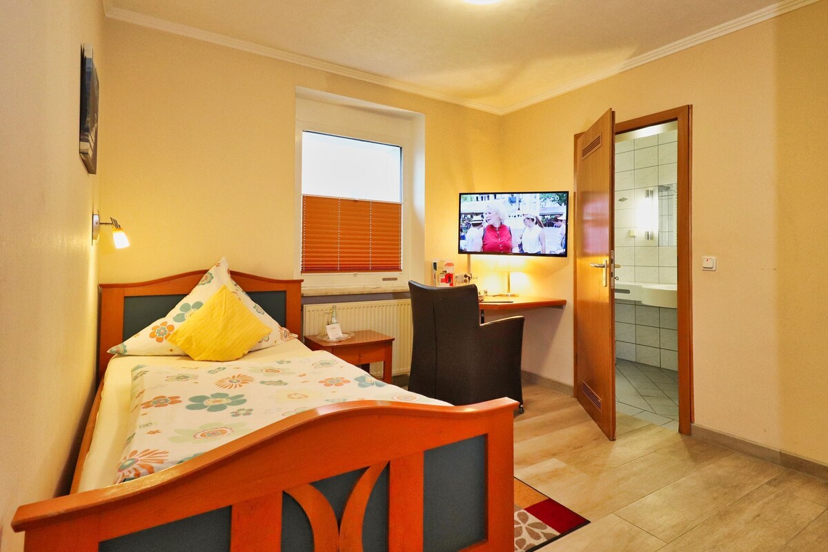 A hotel room with a bed, a desk, a television, and a bathroom visible through an open door.