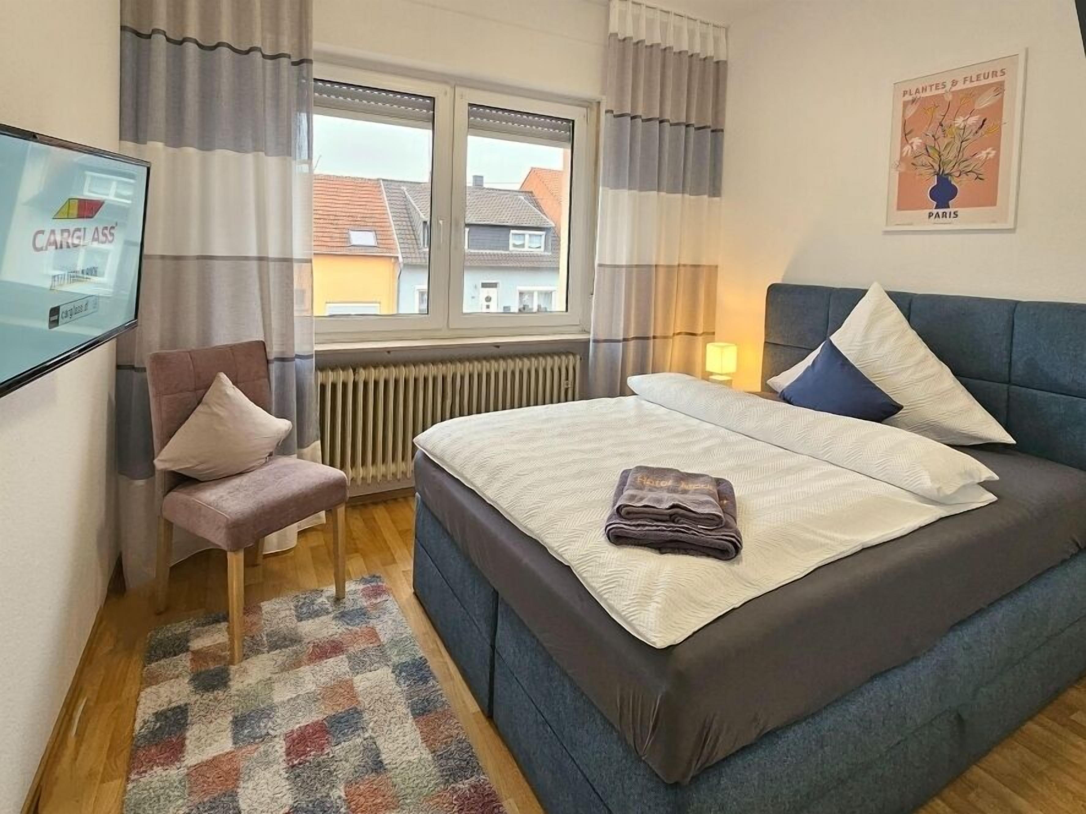 A hotel room with a large bed, a chair, a radiator, a window with curtains, a wall-mounted TV, and a poster on the wall.