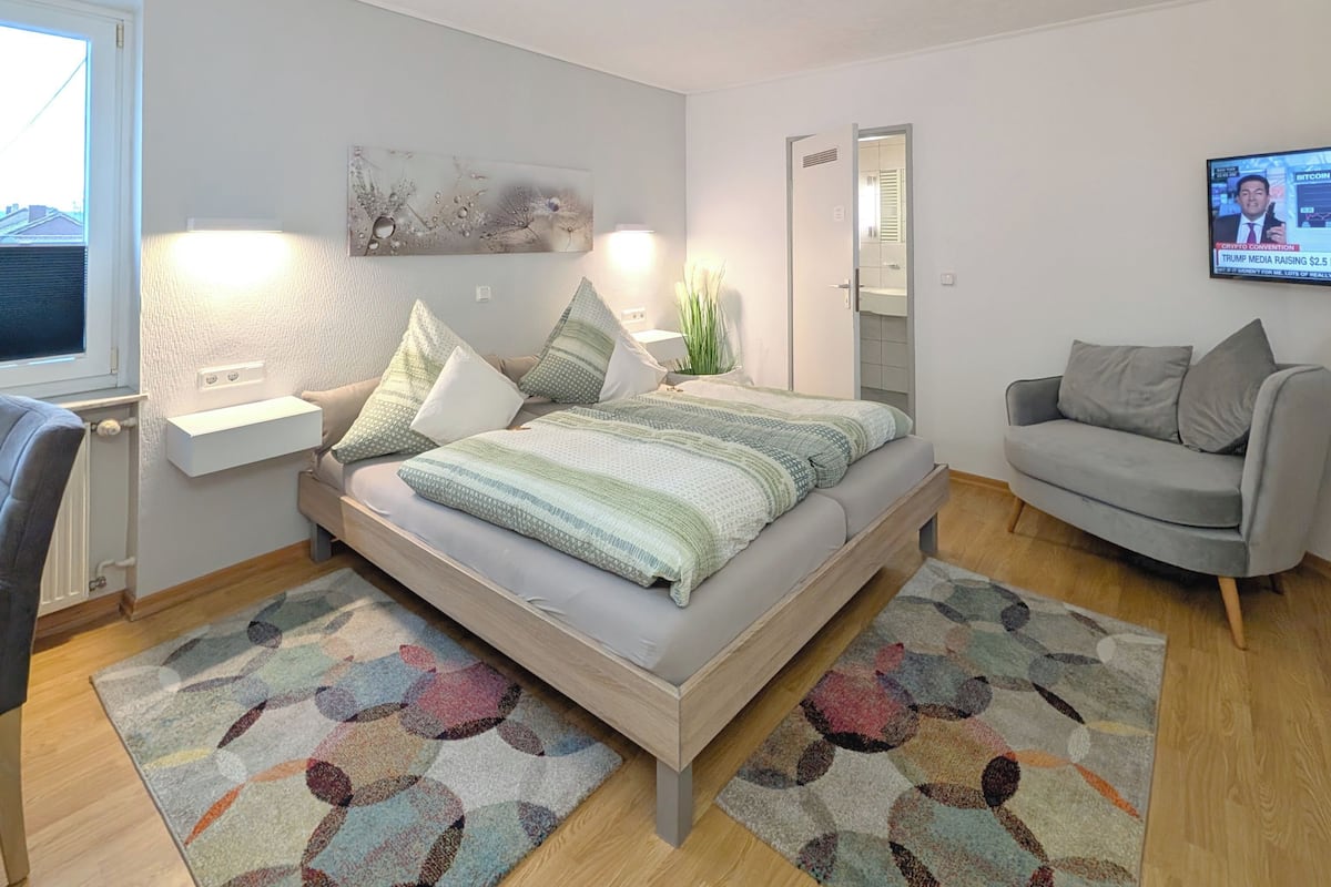 Een moderne slaapkamer met een bed, een grijze bank, een televisie en een schilderij aan de muur.