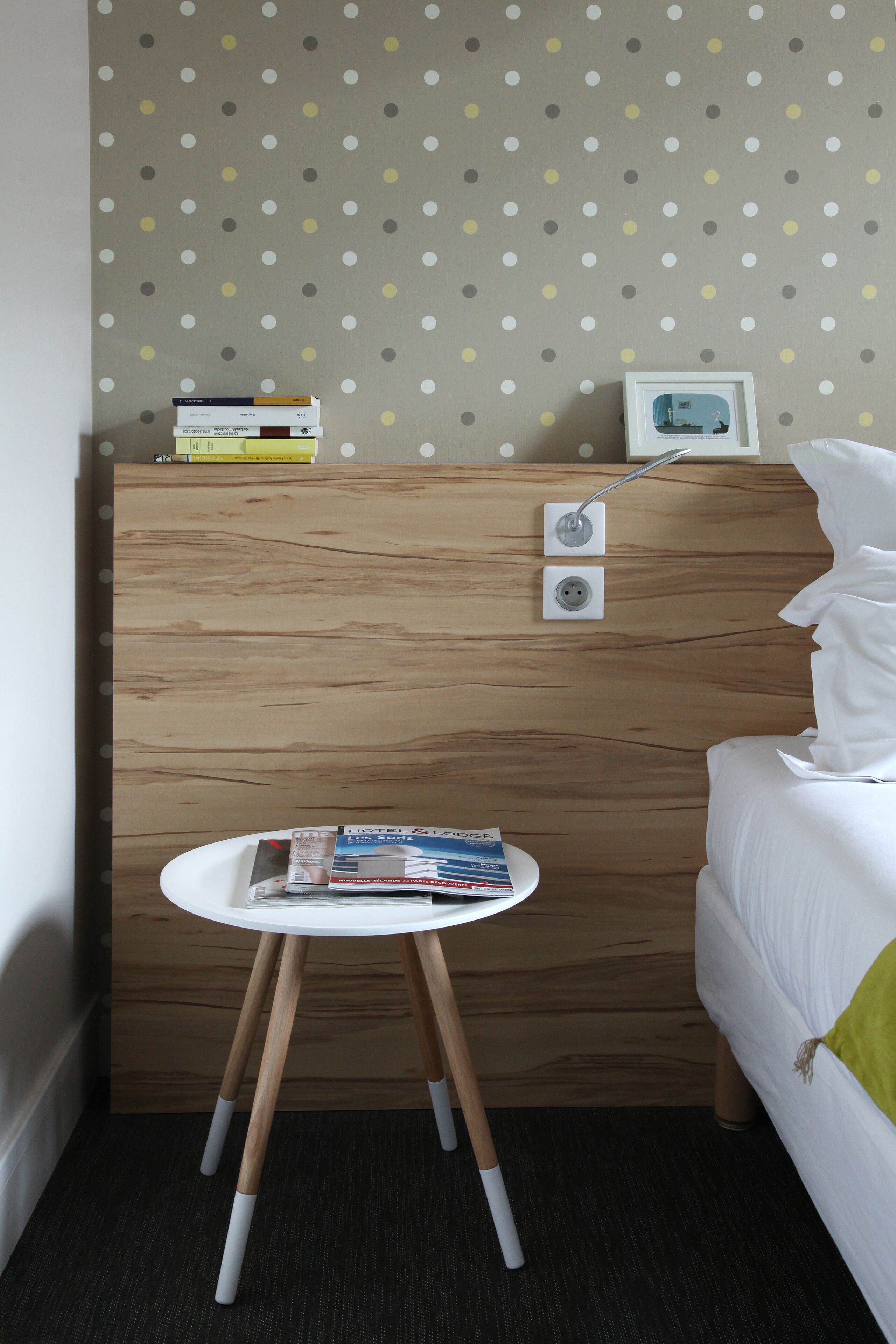 A neatly arranged hotel room with a wooden headboard, a small white table with magazines, and a polka dot wallpaper.