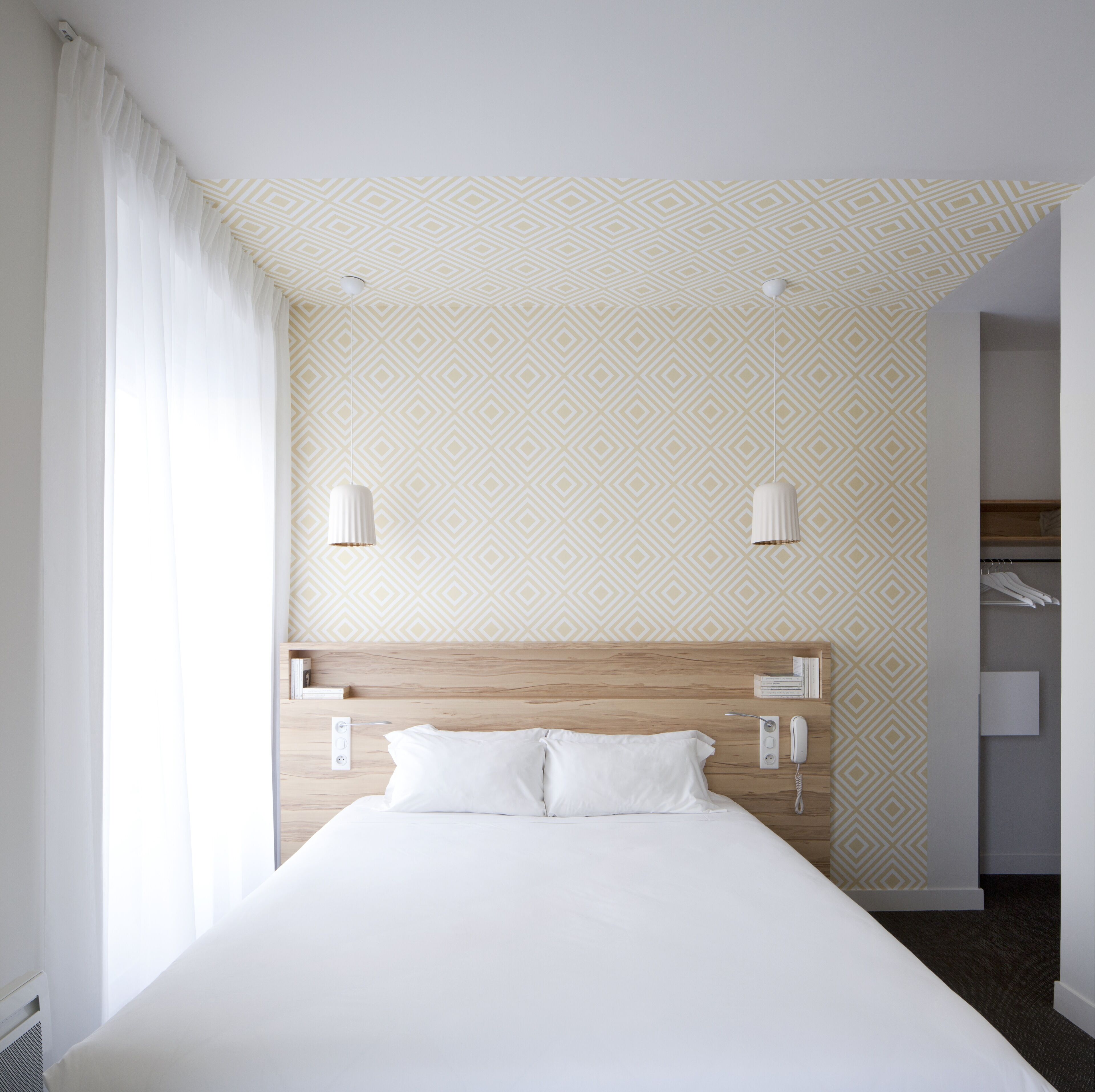 Habitación de hotel con una cama grande, cabecera de madera, pared con estampado, cortina blanca y armario con ropa colgada.