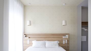 Habitación de hotel con una cama grande, cabecera de madera, pared con estampado, cortina blanca y armario con ropa colgada.