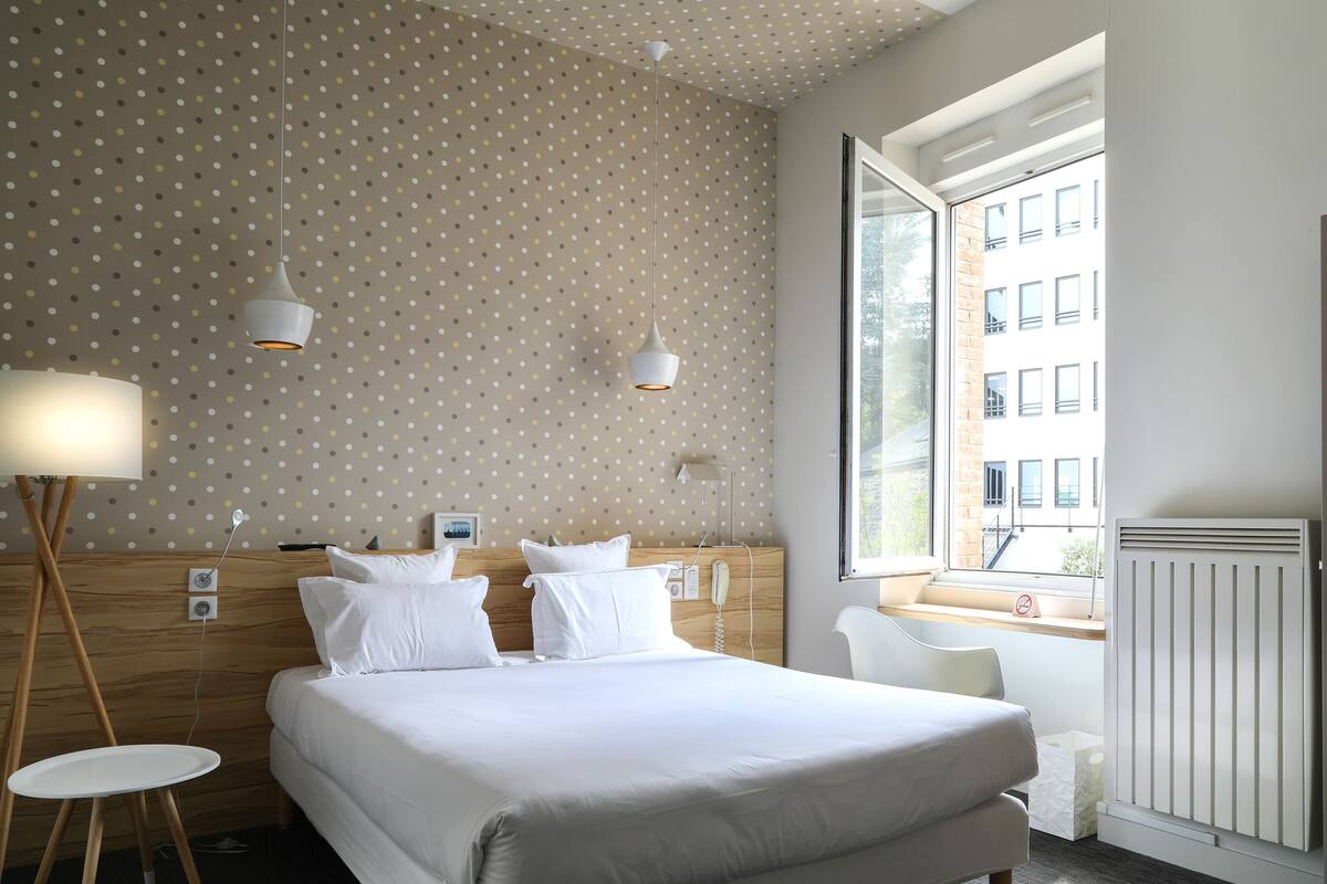 Una habitación de hotel con cama, mesita de noche, lámpara y ventana con vistas a edificios.