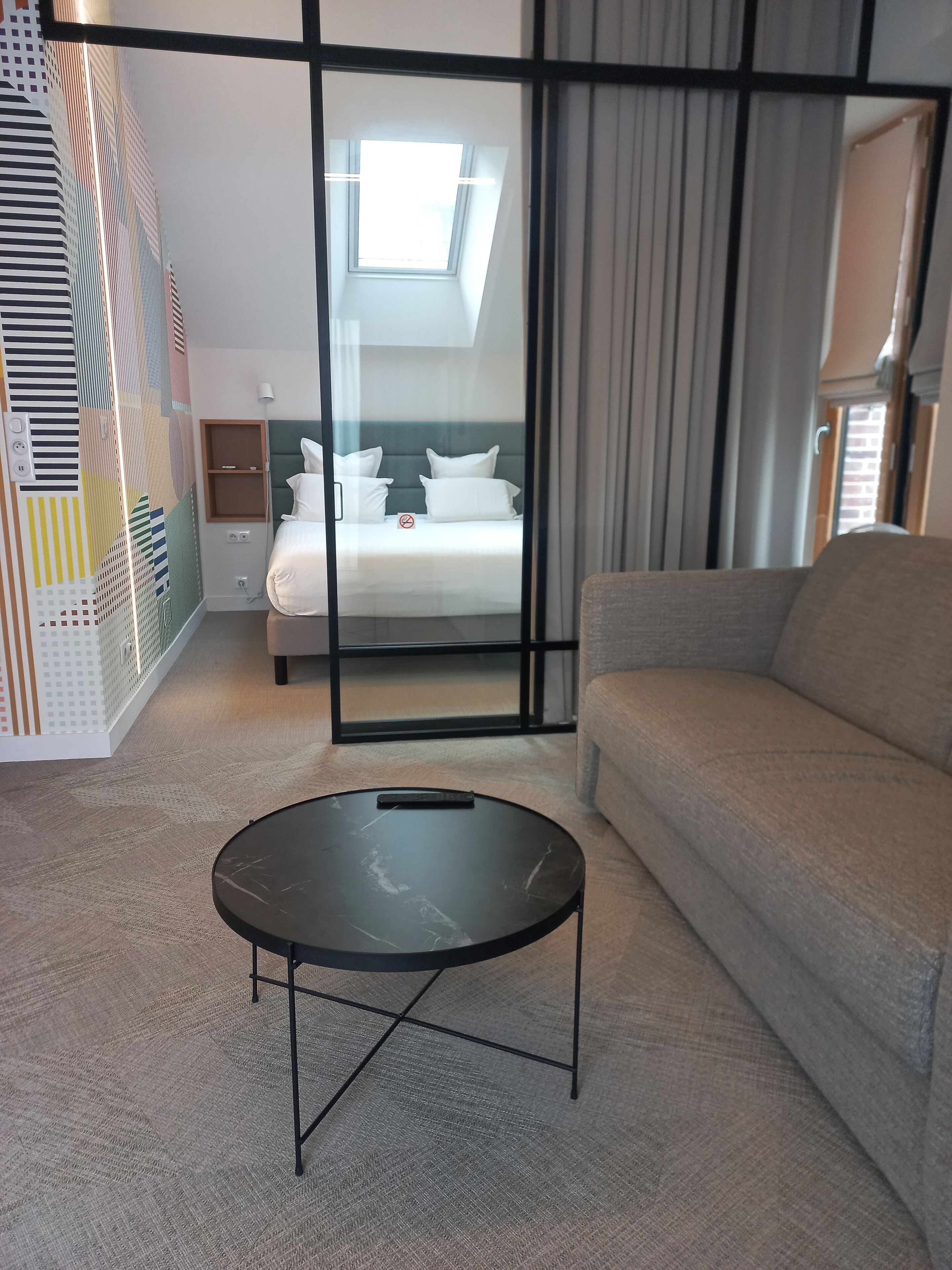 Una habitación de hotel moderna con cama, sofá, una mesita y un ventanal con cortinas.