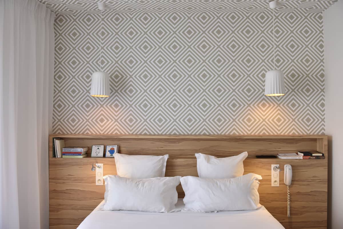 Una cama bien hecha con sábanas y almohadillas blancas, cabecera de madera, teléfono en la mesita de noche y una estantería con libros y una foto enmarco.