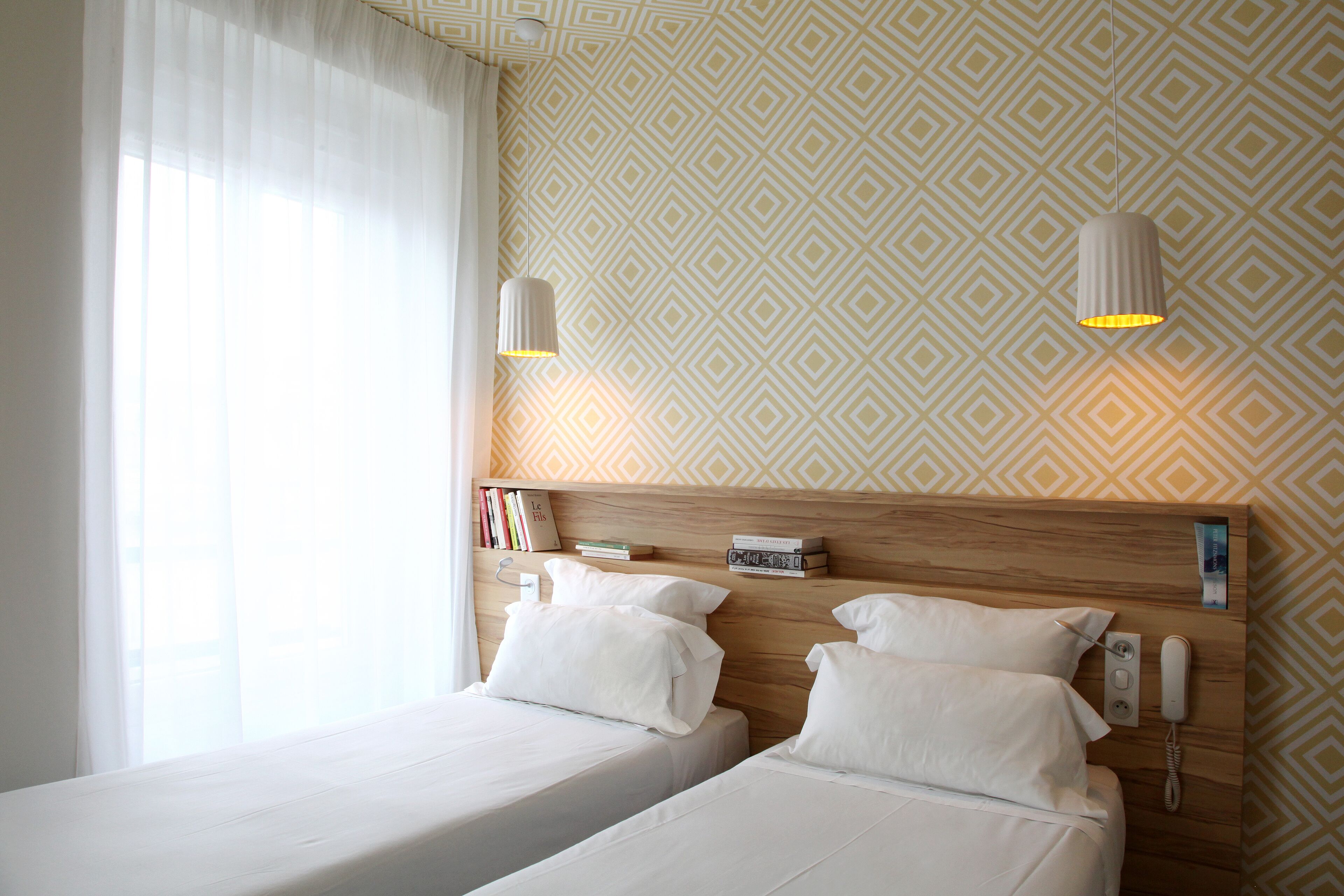 A hotel room with two beds, a wooden headboard, a bedside telephone, and a bookshelf with books.