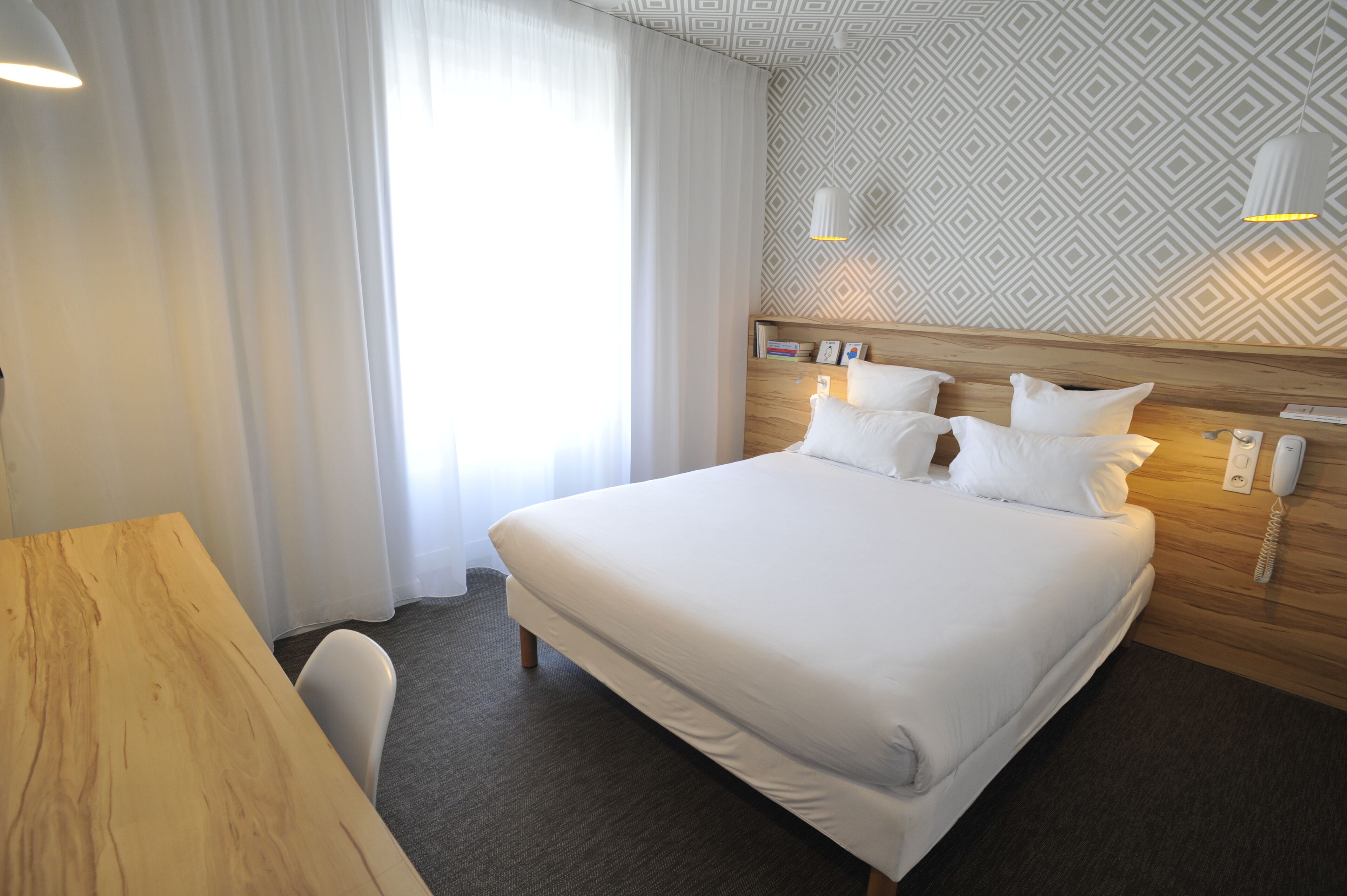 Une chambre d’hôtel avec un grand lit, une tête de lit en bois, un bureau et une chaise. Il y a une fenêtre avec des rideaux, un mur à motifs et un téléphone fixé au mur.