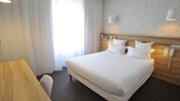 Une chambre d’hôtel avec un grand lit, une tête de lit en bois, un bureau et une chaise. Il y a une fenêtre avec des rideaux, un mur à motifs et un téléphone fixé au mur.