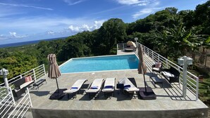 A poolside area with lounge chairs and umbrellas, surrounded by lush greenery and a clear view of the ocean.