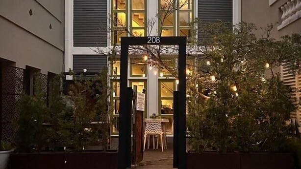 Une entrée d’immeuble avec une plaque portant le numéro 208, un espace extérieur abrité avec des sièges, et une vue sur l’intérieur à travers les fenêtres.