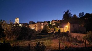 A historic village at dusk with stone buildings, a prominent tower, and a lit-up house. - The house under the stars (Sembadel)