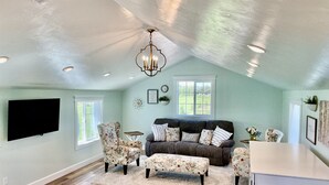 A spacious living room with a high ceiling, a large window, a flat-screen TV mounted on the wall, a dark-colored sofa with floral cushions, and a wooden floor. - The Garden Loft at Heavensfield (Rexburg)