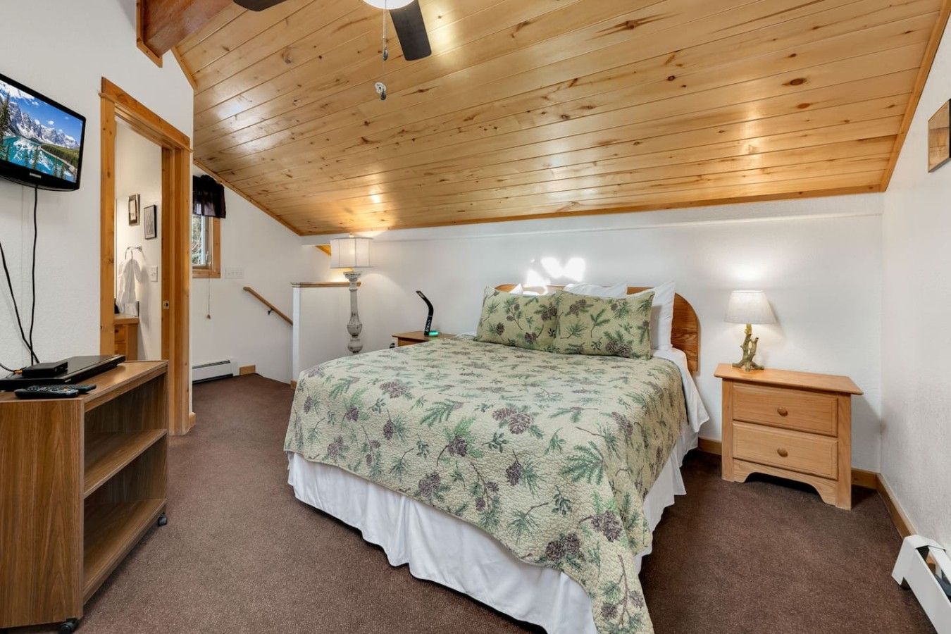Un dormitorio con techo de madera, una cama con ropa de cama floral, una mesita de noche con una lámpara, un escritorio con una computadora y un televisor en la pared.