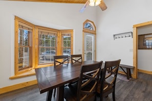 A dining area with a wooden table and chairs, large windows with blinds, a ceiling fan, and a wall-mounted coat rack. - Fall River Oasis • Private Hot Tub • RMNP (Estes Park)