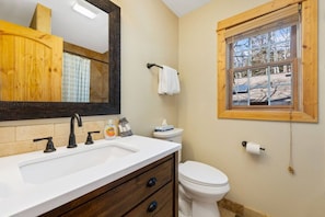 Une salle de bain avec un lavabo blanc, un robinet noir, un meuble en bois et une fenêtre donnant sur des arbres.