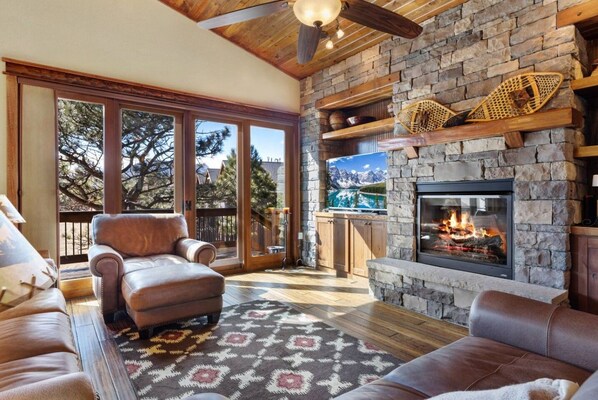 A cozy living room with a stone fireplace, leather sofas, and a large window overlooking a mountainous landscape.