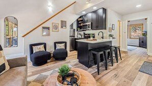 A modern kitchen with dark cabinets, a central island, and bar stools. There are two black armchairs and a sofa in the living area. - Fall River Suite • River Views • Near RMNP (Estes Park)