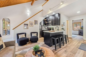 Una cocina moderna con armarios oscuros, una isla central y taburetes de bar. Hay una zona de estar con dos sofás y una mesa de centro.