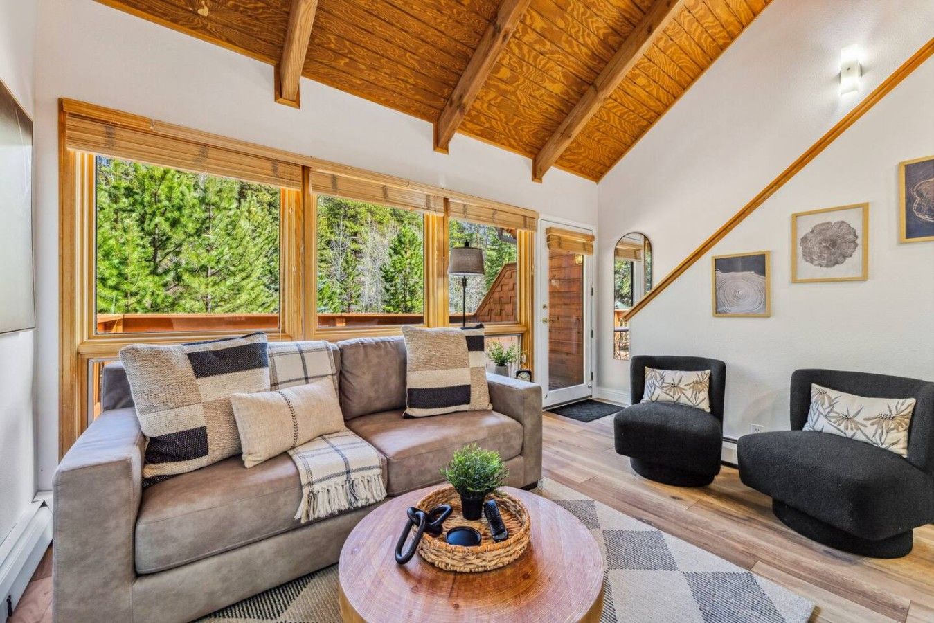 Una sala de estar acogedora con un sofá, dos sillones, una mesita de centro y ventanas amplias con vistas a  árboles.