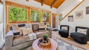 A cozy living room with a sofa, two armchairs, a coffee table, and large windows offering a view of trees. - Fall River Suite • River Views • Near RMNP (Estes Park)