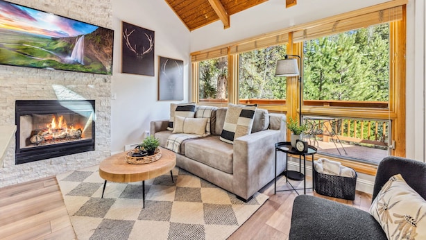 A cozy living room with a fireplace, a sofa, a coffee table, and large windows offering a view of the outdoors. - Fall River Suite • River Views • Near RMNP (Estes Park)