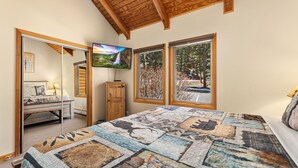 A bedroom with a large bed, a TV on the wall, and a view of trees through the windows. - Fall River Suite • Serene • Close to RMNP (Estes Park)