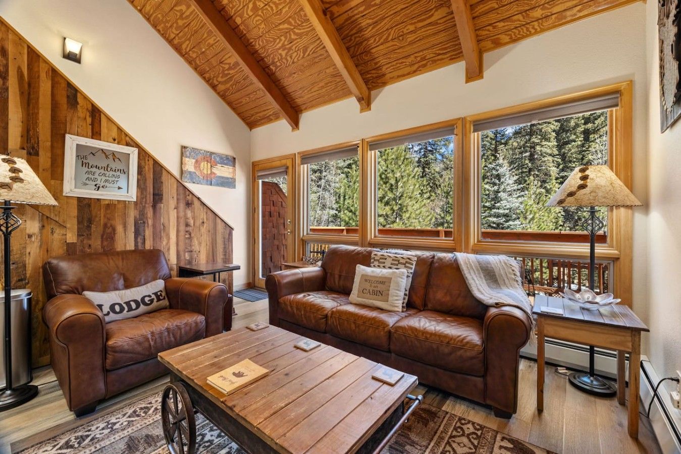 Interior acogedor de una cabaña con techo de madera, muebles de cuero y ventanas amplias que dan a un bosque.