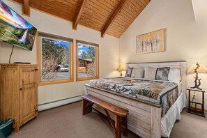 A bedroom with a large bed, wooden headboard, two windows, a bench, a cabinet, and a painting.
