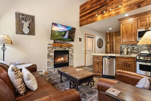 A living room with a fireplace, a television mounted above it, and a kitchen area with wooden cabinets and appliances.