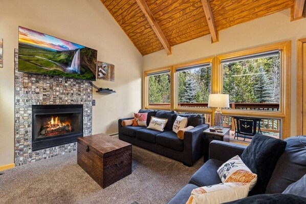 A cozy living room with a fireplace, a large window with a view of trees, and a comfortable seating area.
