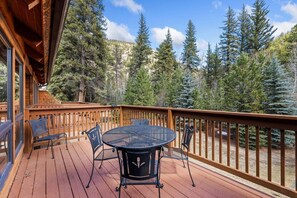 A wooden deck with a round table and chairs, surrounded by a wooden railing, overlooking a forested area.