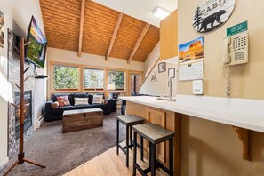A cozy living area with a fireplace, a sofa with cushions, a coffee table, and a kitchen area with a counter and stools.