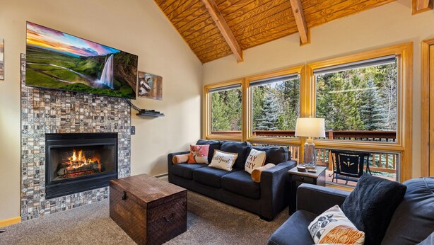 A cozy living room with a fireplace, comfortable seating, large windows, and a wooden ceiling.