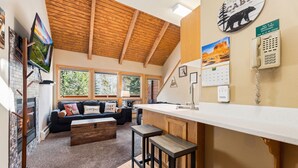 A modern kitchen with a wooden ceiling, a white countertop, and a bar area. There is a large window with a view of trees and a fireplace.