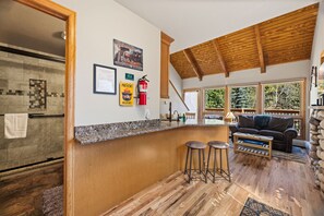 A kitchen with a wooden ceiling, a bar area with stools, a fire extinguisher, and a view of trees through large windows. - Peaceful Fall River Suite • Fireplace• Close to RMNP (Estes Park)