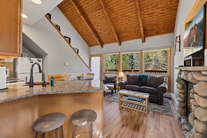 A modern kitchen with a wooden ceiling, a bar area with stools, a fireplace, and large windows offering a view of the outdoors. - Peaceful Fall River Suite • Fireplace• Close to RMNP (Estes Park)