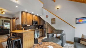 A modern kitchen with wooden cabinets, a central island, bar stools, and a dining area with a round table. - Riverfront Haven • River Views • Near RMNP (Estes Park)