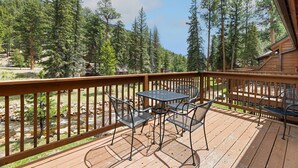 A wooden deck with a table and chairs, surrounded by a wooden railing, overlooking a forested area. - Riverfront Haven • River Views • Near RMNP (Estes Park)