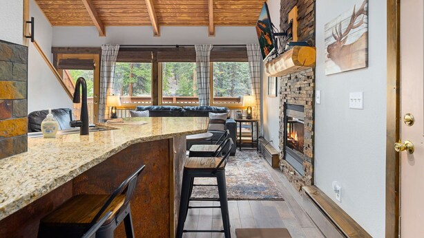 A modern kitchen with a stone fireplace, wooden ceiling beams, and a bar area with stools. - Riverfront Haven • River Views • Near RMNP (Estes Park)