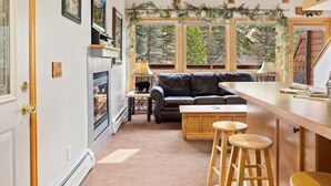 Ein gemütliches Interieur mit Kamin, einer Ledersofa und hölzernen Barhocker, sowie großen Fenstern mit Blick ins Freie.
