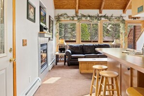 Un intérieur chaleureux avec une cheminée, un canapé en cuir, un coin bar en bois et de grandes fenêtres offrant une vue sur l’extérieur.