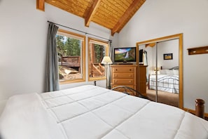 A bedroom with a large bed, wooden ceiling, a television, a dresser, and a mirror reflecting a bed. - Cozy Riverfront Suite • Near RMNP • River Views (Estes Park)