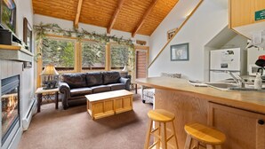 Ein gemütliches Wohnzimmer mit Kamin, einer Couch und einem Couchtisch. Es gibt eine Küchenzeile mit Kühlschrank und Spüle.