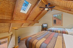 Ein Schlafzimmer mit schrägen Wänden, einem Deckenventilator, einem Fernseher auf einem kleinen Tisch und einem Fenster mit Blick auf ein Gebäude.