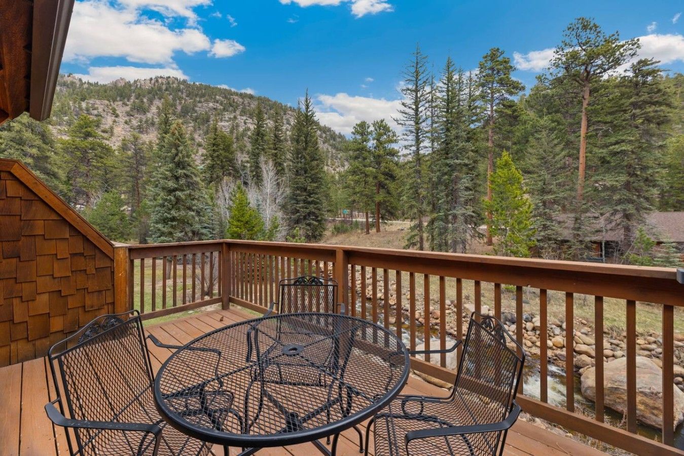 Una terraza de madera con una mesa y sillas de metal, con vistas a un paisaje montañoso y árboles.
