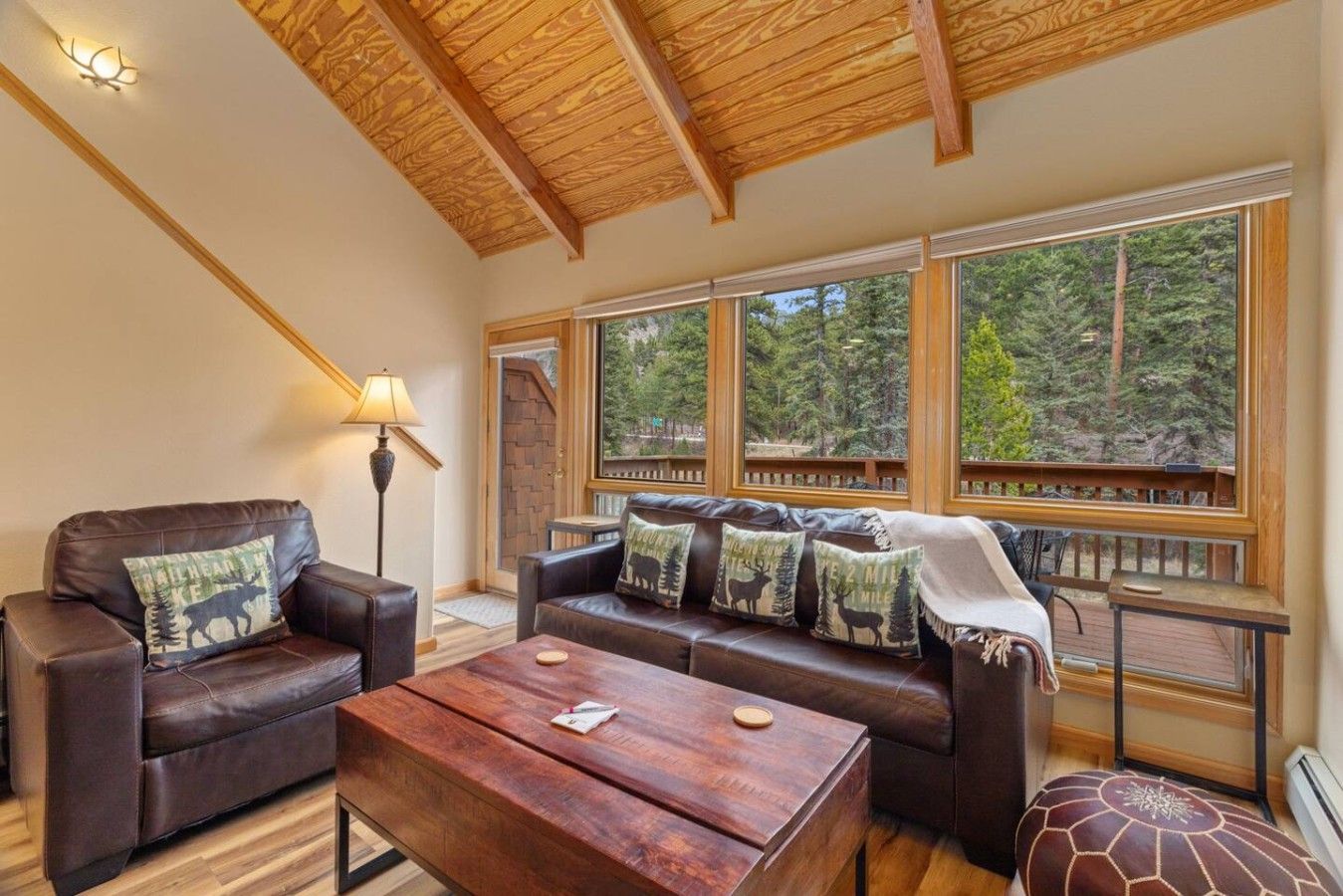 Una sala de estar acogedora con techo de madera, un sofá de cuero con cojines decorativos, una mesa de centro de madera y grandes ventanales con vistas al bosque.