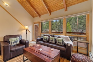 Una sala de estar acogedora con techo de madera, un sofá de cuero con cojines decorativos, una mesa de centro de madera y grandes ventanales con vistas al bosque.
