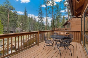 A wooden deck with a table and chairs overlooking a forested area and a stream. - Quiet Fall River Suite • Fireplace • Close to RMNP (Estes Park)