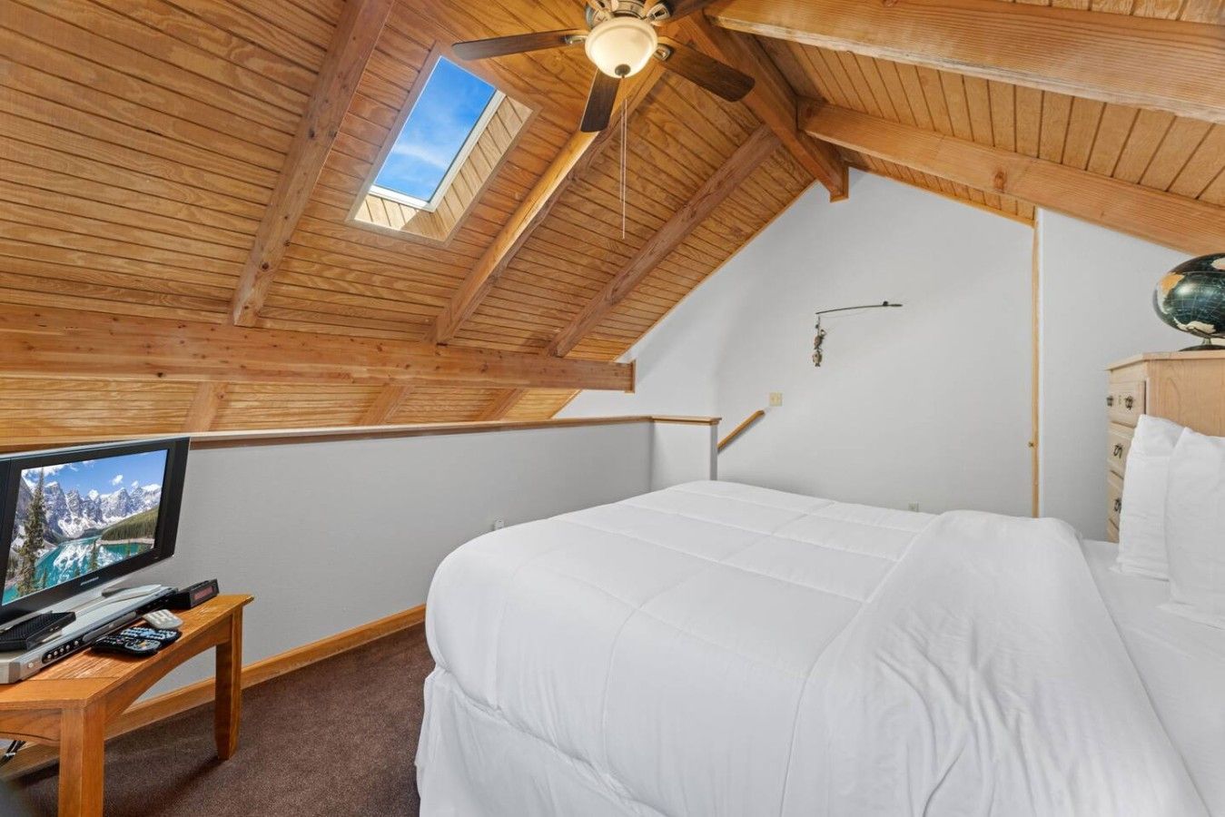 Un dormitorio con techo inclinado, una cama, un televisor sobre una mesa de madera y un ventilador de techo.