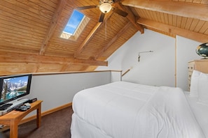 Une chambre avec un plafond en pente, un lit, un téléviseur posé sur une table en bois et un ventilateur de plafond.